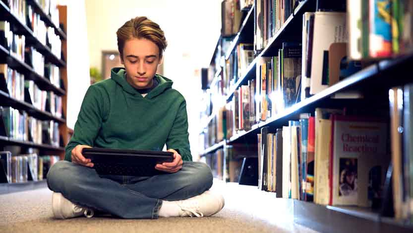 A boy sitting on the floor in a library and looking at his tablet