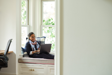 Image of a smiling woman with glasses working on a laptop computer in a cushioned bay window. She is laid back against a big pillow and there is foliage outside the window.