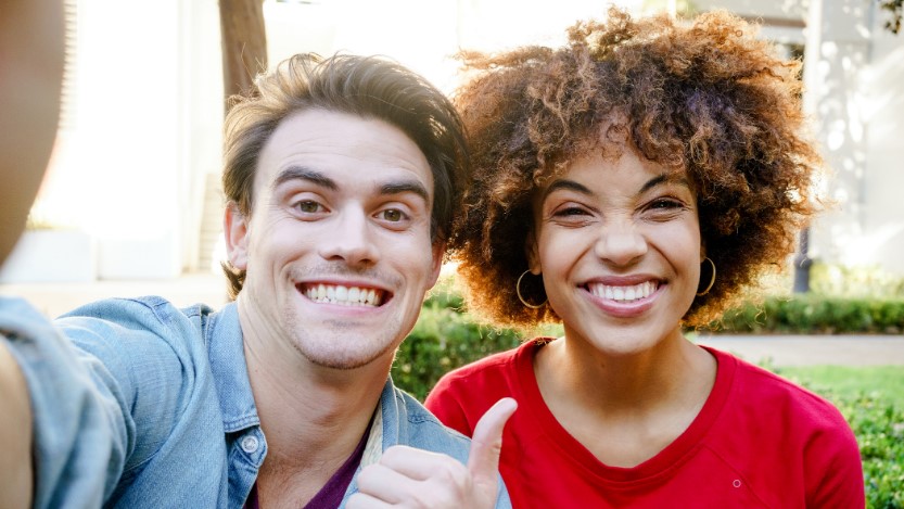 Selfie of male and female friends in outdoor park. Both are smiling and looking at camera. The man is making a thumbs up sign on left hand, with his right arm extended toward camera.