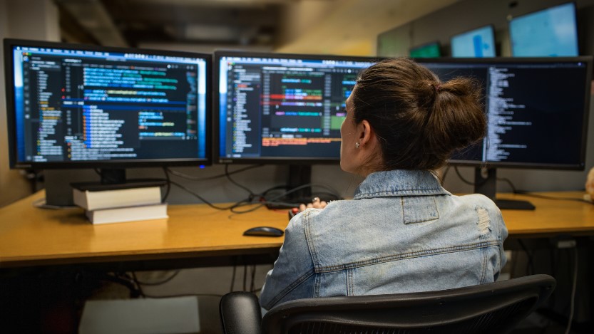 Woman sitting at a desk at work, looking at three monitors and running an analysis.