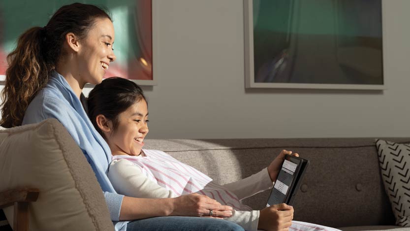 A parent and child sitting on a couch smiling while they look at a tablet screen. 