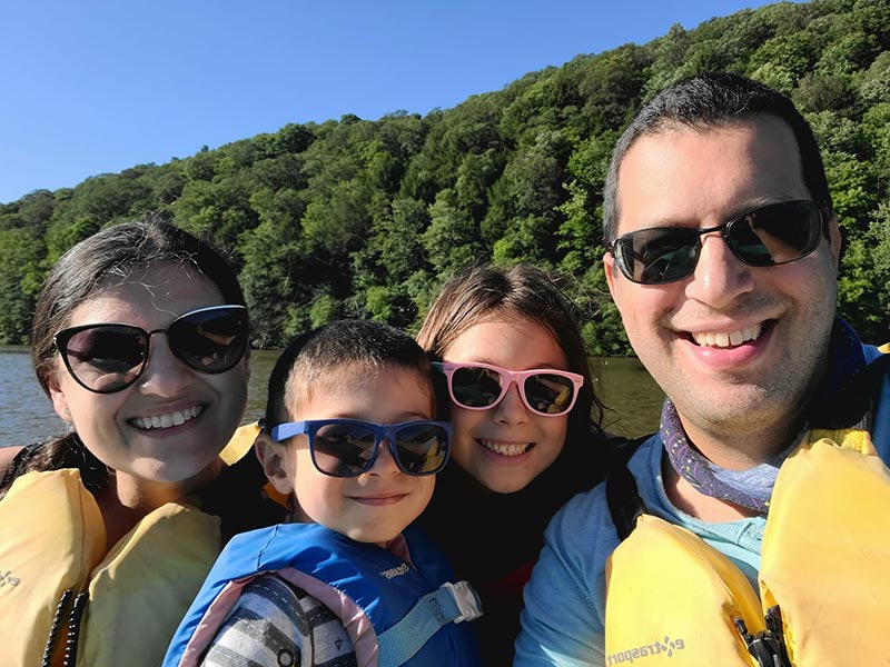 Digital nomad Audrey Frankel and her family, wearing flotation vests by a lake on a sunny day, smile at the camera.