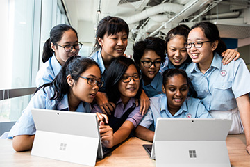 A group of girls sit and stand in front of two Microsoft tablets.