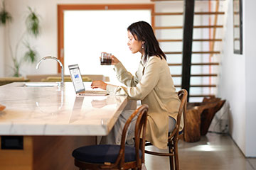 A woman sits in her kitchen sipping coffee and working on her Microsoft Surface laptop PC.