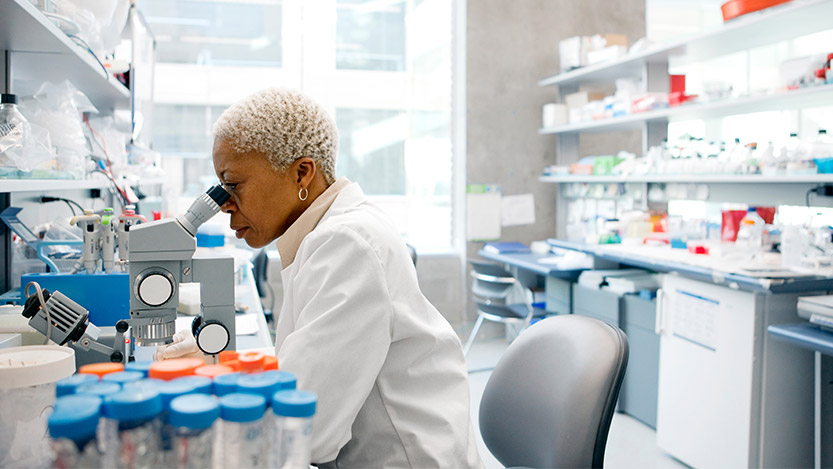 A woman with a white lab coat sits in a lab doing research with a microscope.