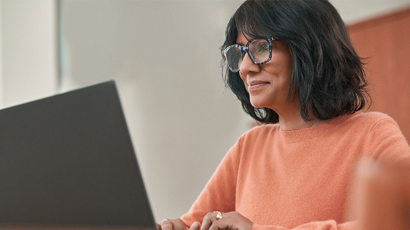 A woman with glasses slightly smiles while looking at a computer.