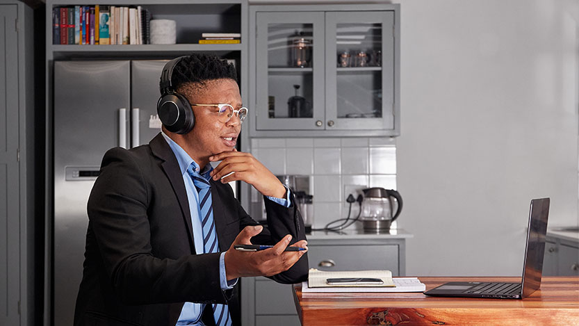 A man wearing headphones is sitting in a kitchen while attending a meeting on a laptop.