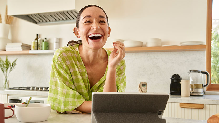 A woman sitting in a kitchen laughs while using her tablet.