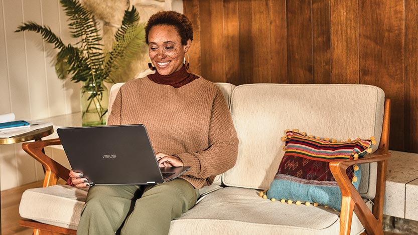 A woman sitting on a couch smiles while working on a laptop.