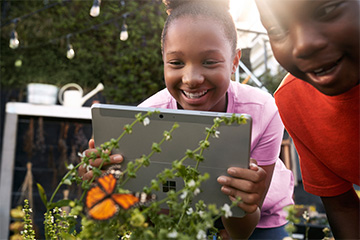 Two kids outdoors photograph a butterfly with their tablet.