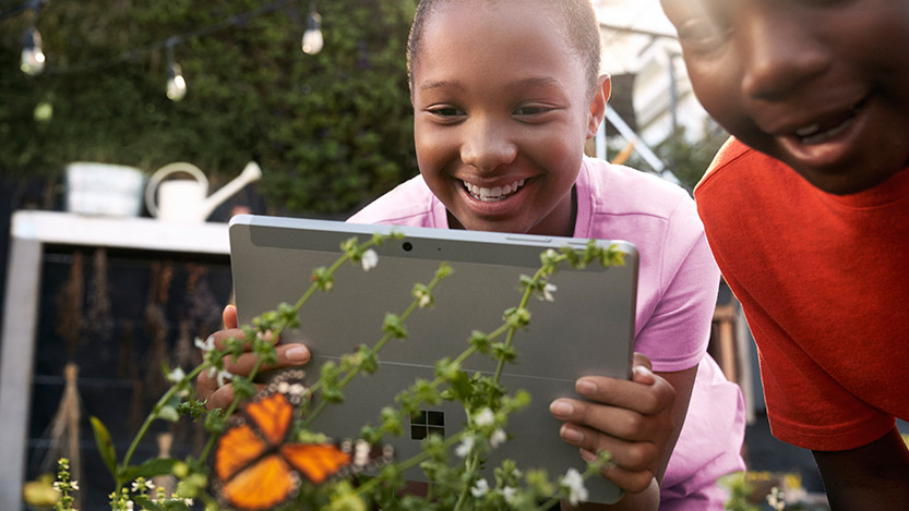 Two kids outdoors photograph a butterfly with their tablet.