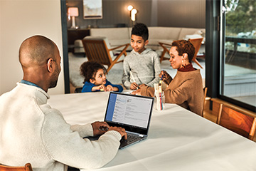 A man works on his computer while a woman and two children sit across a table.