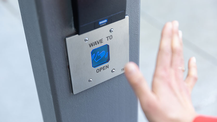 A hand moves in front of a digital door system that asks you to “wave to open”.