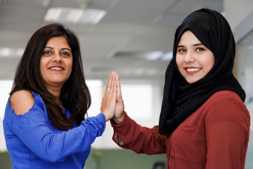 Two smiling female workers high fiving in workplace. The woman at right wears a headscarf; the woman at left wears a long sleeve sweater with open shoulders.