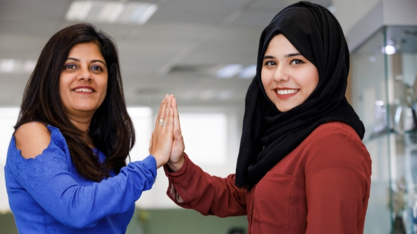 Two smiling female workers high fiving in workplace. The woman at right wears a headscarf; the woman at left wears a long sleeve sweater with open shoulders.