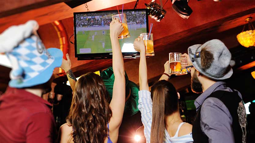 A group of people at a bar watching a football match celebrates by raising their glasses