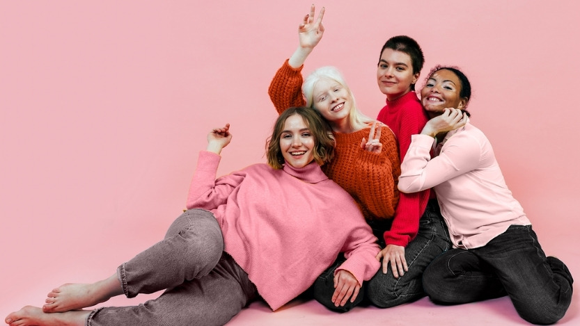 Group of diverse young women wearing shades of red and pink, smiling directly at the camera relaxing on the ground.