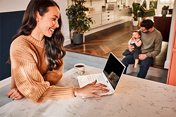 A woman smiles while working on a laptop computer while a man and a baby are interacting in the background