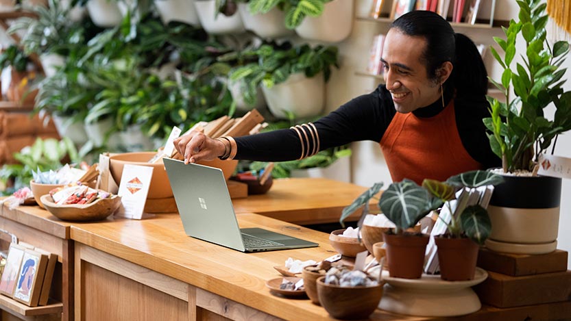 A man inside a shop surrounded by plants works on a laptop while smiling.