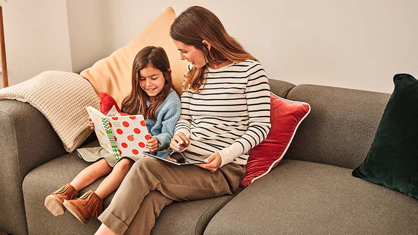 A mother and her daughter reading a book on the couch at home.