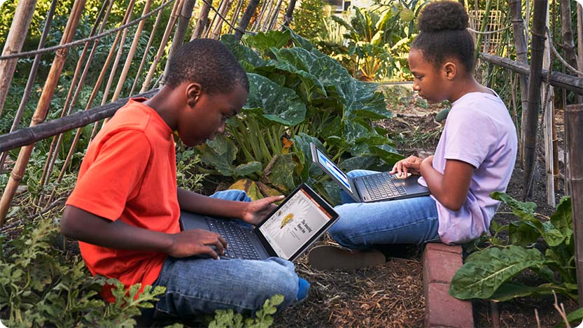Two kids sitting in a garden, both with laptops.