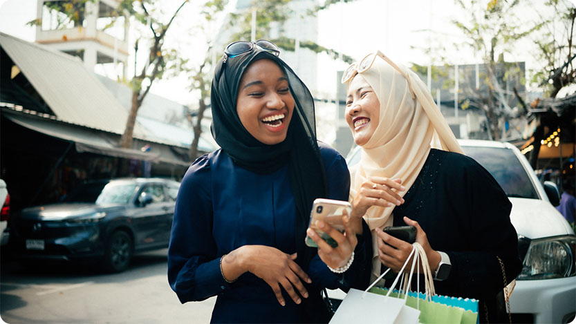 Two people laughing on the street while holding shopping bags and phones.