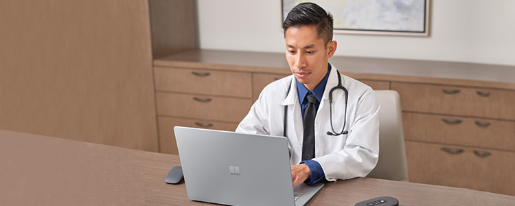 An Asian man in a white lab coat sits at a table in an office, typing on a Microsoft Surface laptop PC.