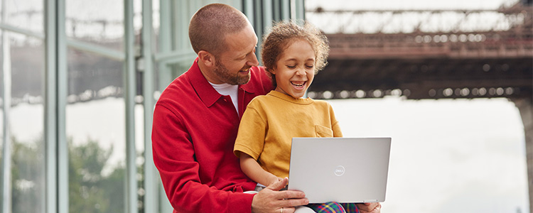 A girl sits on her father's lap while laughing and looking at a laptop.