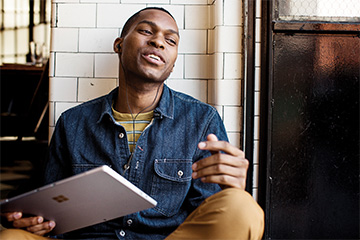 A young man sits against a wall while listening to something on his Microsoft tablet.