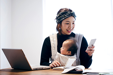 A woman sitting and working with her laptop holds a baby in a carrier and smiles while looking at her phone.