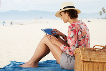 A woman with a hat uses a tablet while sitting on the beach.