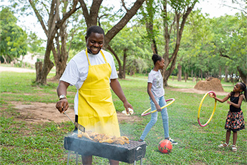 A man wearing an apron prepares a BBQ while two young girls play with hula hoops behind him.
