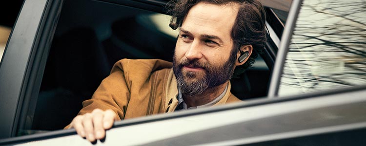 A man wearing earbuds looks away from the camera and slightly smiles while getting out of a car.