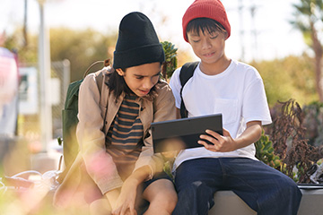 Two children sitting outside look at a tablet.