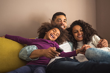 A family of three smiling and cozying up on a couch together while watching TV.