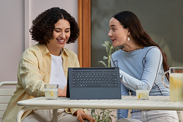 Two women in a casual setting sit at a table with glasses of lemonade and laugh while watching something on their laptop.