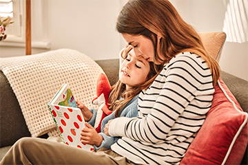 A woman leans her head on a young girl’s while they sit on a couch and read a children’s book.