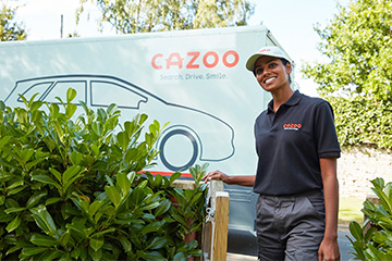 A woman with a Cazoo uniform and hat stands in a driveway with a Cazoo truck in the background.