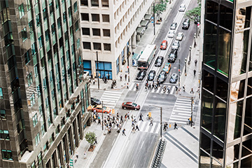 A bird's view of an intersection in a city, surrounded by buildings and with cars and buses waiting to move.