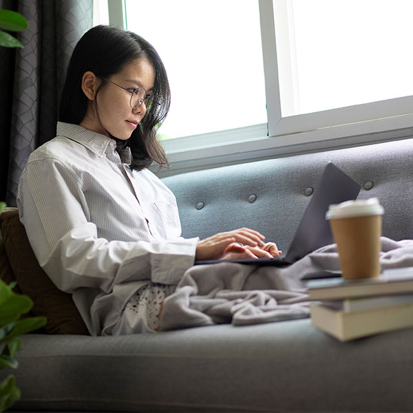 ソファに座って足を伸ばし、ノートパソコンで作業をしている若い女性。