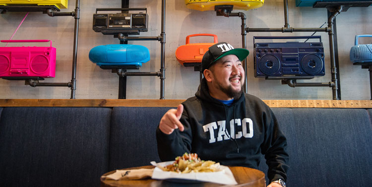 At a modern restaurant, a man with a hat on sits on a bench and points at a plate with tacos while smiling away from the camera.
