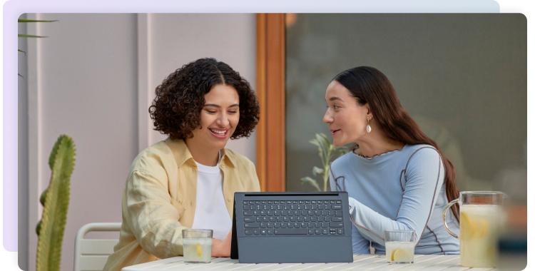 Two women talk while sitting at a table and working on a laptop computer.