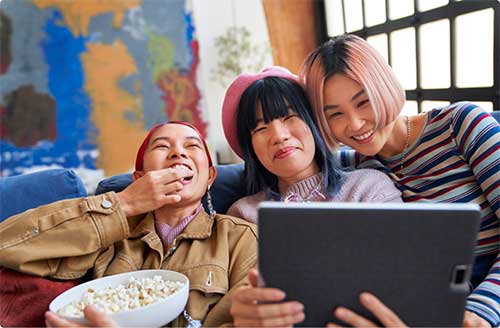 Three women sit in front of a tablet while eating popcorn.