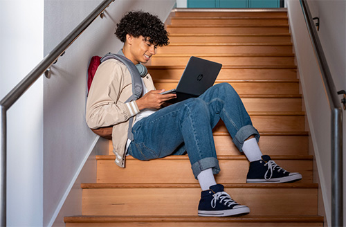 A young man sits on the stairs while working on his laptop computer.