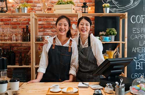 Two women wearing aprons smile behind the counter of a coffee shop and smile at the camera.