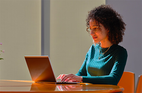 A girl wearing glasses sits at a table and uses her laptop.
