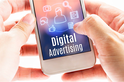 A close-up of hands holding a cellphone in which the screen shows a person icon surrounded by other media-related icons. The text below reads: “Digital Advertising”.