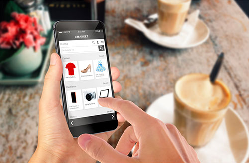 Two hands hold a smartphone while navigating through a catalog with items from apparel to technology. A wooden table and two iced coffees can be seen in the back.