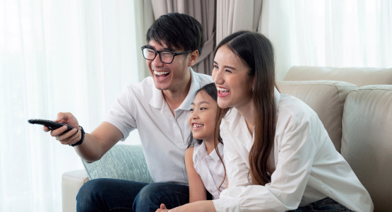 Two adults and a child watch TV and laugh while sitting in a couch.