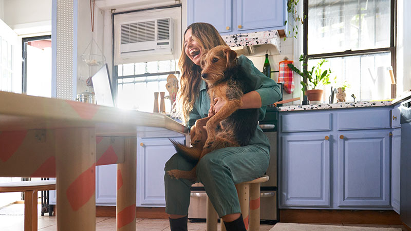 In a home kitchen, a women holds her dog and laughs at something she sees on her laptop screen.